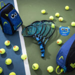 Três raquetes de tênis infantis pretas e azuis da linha Head Novak empilhadas no centro de uma quadra com piso verde e azul. Ao redor das raquetes, há várias bolas de tênis espalhadas, uma mochila infantil azul com o desenho de um rosto de monstrinho, uma raqueteira azul e preta, e uma pequena máscara de papelão de monstro com a marca Head